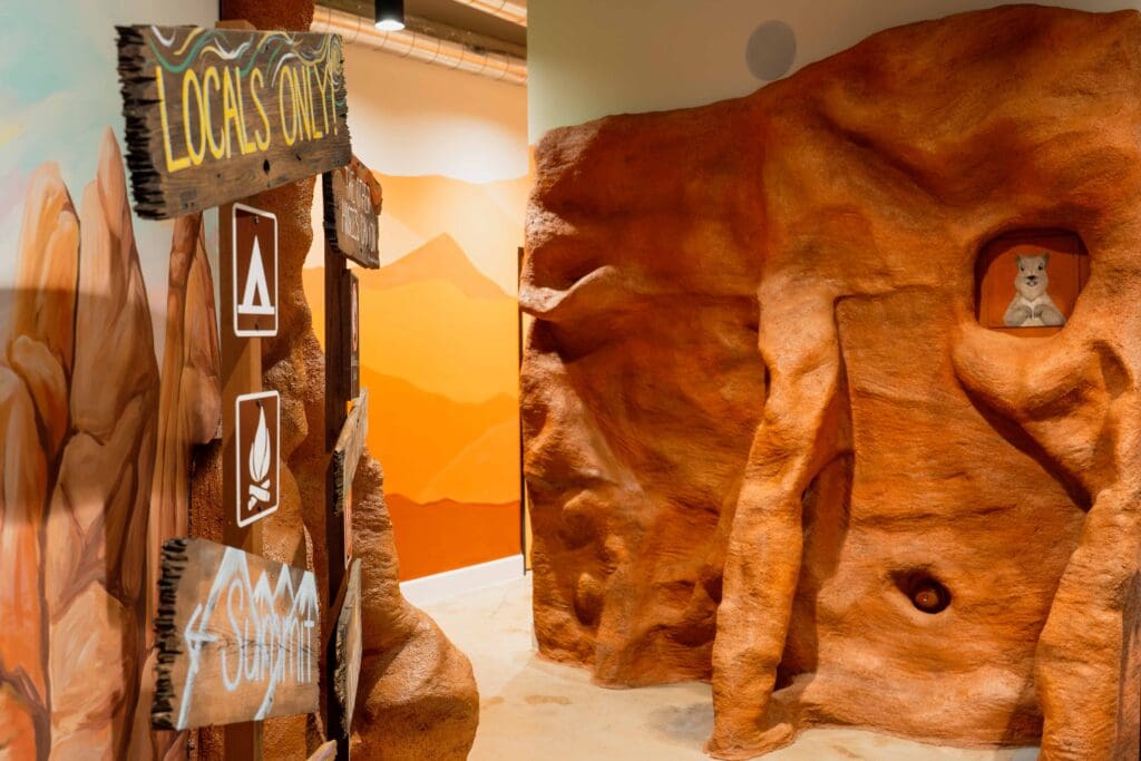 Biophilic hospitality hallway inspired by sand canyons, featuring sculptural rock-textured walls, warm earth tones, and a nature-inspired path leading to the restrooms at Bivouac Adventure Lodge.