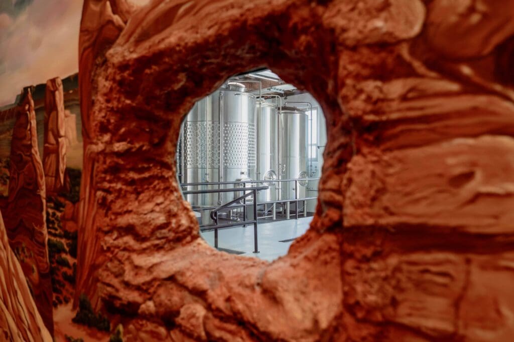 Biophilic hospitality design detail showing a rock formation mural with a circular peep-hole window revealing the production space beyond, creating a nature-inspired moment of curiosity and connection.