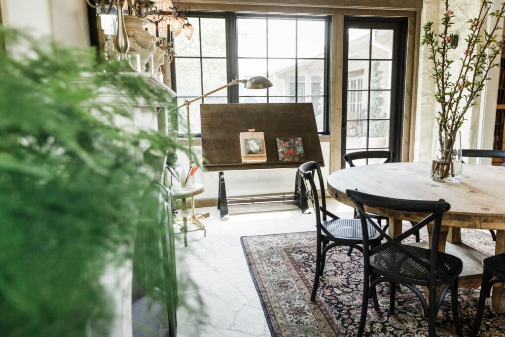 Sunlit creative studio with drafting table, round wooden table, black chairs, large windows, and natural greenery creating a calm, intentional workspace.